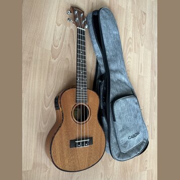 Dřevěný ukulele v hnědé barvě s přiloženým šedým textilem obalem. Na hlavě nástroje i na obalu je bílý nápis CASCHA. Nástroj má v těle zabudovaný pre…