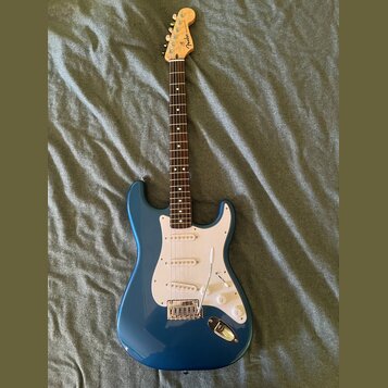 Modrá elektrická kytara s bílým pickguardem a světle hnědým hmatníkem. Na hlavě je viditelné logo Fender. Nástroj má tři snímače, bílé ovládací knofl…