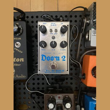 Stříbrný efekt pedál s nápisy „OUT“, „9V DC“, „IN“, „vol“, „gain“, „bass“, „mid“ a „treble“. Uprostřed je modrý nápis „welcome to ,you're Doom 2“ a v…