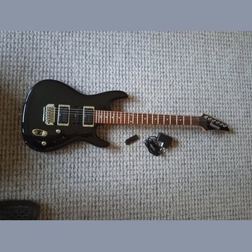 Černá elektrická kytara s tmavým hnedým hmatníkem a dvěma humbuckery. Na hlavě nástroje je bílý nápis Ibanez. Vedle kytary leží černý napájecí adapté…