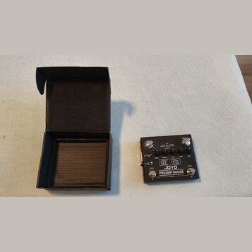 Černý kytarový předzesilovač s nápisy JOYO a PREAMP HOUSE. Na horní straně jsou čtyři otočné ovladače, přepínač a několik textových popisků včetně BA…