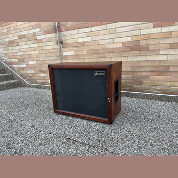 Hnědý basový reproduktor v dřevěném korpusu s černou textilní maskou. V pravém horním rohu masky je umístěn štítek s nápisem „gbr Custom Cabinets“. P…