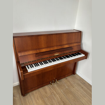 Hnědé dřevěné piano s otevřenou klaviaturou. Na přihradě nad klávesami je zlatý nápis „Behringer“. Nástroj vykazuje známky běžného používání, povrch…
