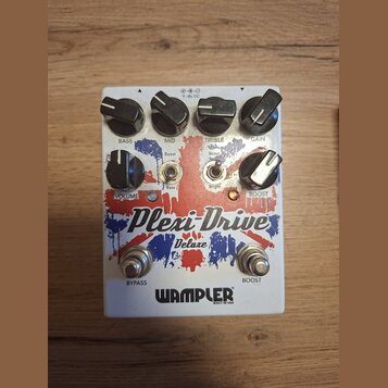 Bílý kytarový efekt s červenými a modrými grafickými prvky. Na zařízení jsou nápisy Plexi-Drive Deluxe a WAMPLER BUILT IN USA. Horní řada ovládacích…