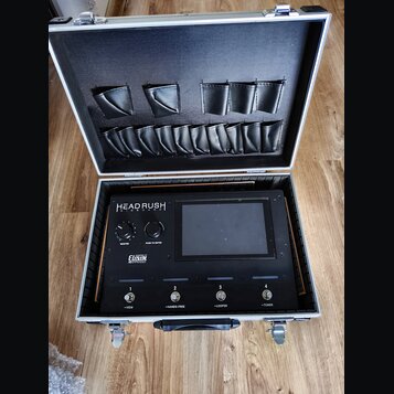 V otevřeném hliníkovém kufru s černým vnitřním polstrováním je umístěno zařízení s nápisy HEAD RUSH GIGBOARD a ELEVEN HD EXPANDED. Přístroj má černý…