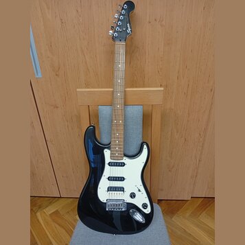Černá elektrická kytara s bílým pickguardem a hnedým hmatníkem. Na hlavě je viditelné logo Squier. Nástroj má tři snímače typu single-coil a chromova…