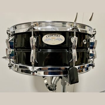 Černý buben s chromovaným obručováním a mechanismy. Na korpusu je oválný štítek s nápisy CONCERT, YAMAHA a JAPAN. Povrch nástroje je glossy, v odrazu…