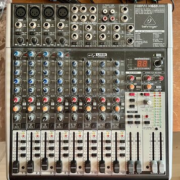 Stříbro-šedý mixážní pult s černými ovládacími prvky. V pravém horním rohu je nápis XENYX X1622USB a logo behringer. Přístroj obsahuje více vstupních…