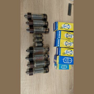 Fotografie zobrazuje skupinu elektronických vakuových elektronek a jejich originálních krabiček na dřevěném povrchu. Na některých elektronkách je čit…