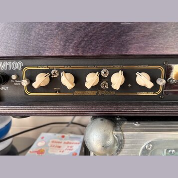 Detail kytarového zesilovače v tmavém dřevěném rámu. Na černém předním panelu jsou bílé oválné knofíky s popisky VOLUME, BASS, MIDDLE, TREBLE a MASTE…