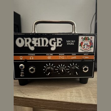 Černý kytarový zesilovač s kovovou rukojetí a oranžovým pruhem na předním panelu. Na horní části je bílý nápis ORANGE a vedle něj text MICRO DARK, v…