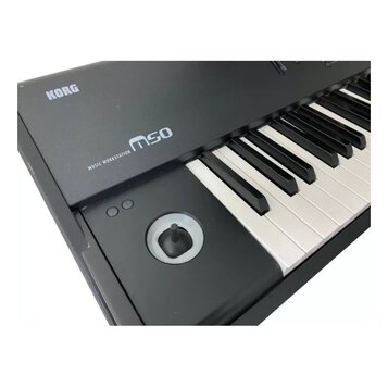 Černý klávesový nástroj s bílými a černými klávesami. Na horním panelu jsou bílé nápisy KORG a MUSIC WORKSTATION m50. V levé části je umístěn kulatý…