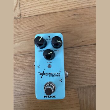 Světle modrý efekt pedál s třemi černými oválnými knoflíky. Na pedálu jsou nápisy SHINE, LEVEL, TONE a DRIVE, dále text MORNING STAR OVERDRIVE a logo…