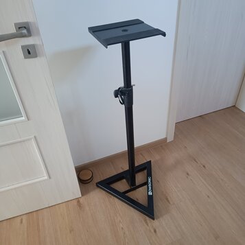 Černý kovový stojan s trojúhelníkovou základnou a plochou horní deskou. Na jedné z hran základny je bílý nápis NOWSONIC. Povrch stojanu vykazuje mírn…