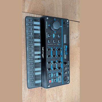 Černý hybridní syntetizátor s modrými detaily a nápisem Pro VS MINI HYBRID VECTOR SYNTHESIZER. Na levé straně je umístěno logo behringer a blok tlačí…