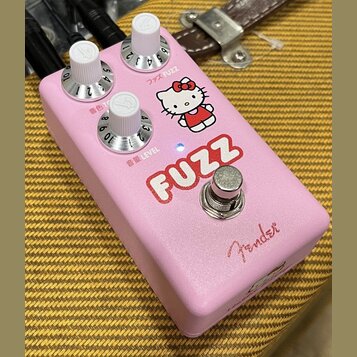 Růžový efekt pedál s nápisem FUZZ a obrázkem postavy Hello Kitty. Na horní části jsou tři bílé ovládací knoflíky s popisky ファズ FUZZ, 音色 TONE a 音量 LEV…