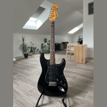 Černá elektrická kytara typu Stratocaster s černým pickguardem a hnedým hmatníkem. Na hlavě je viditelné logo „Fender Stratocaster“. Nástroj je umíst…