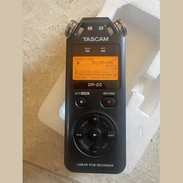 Černý digitální záznamový zařízení s nápisem TASCAM a označením modelu DR-05. Displej je zapnutý s oranžovým podsvícením a zobrazeným textem 00H00M00…