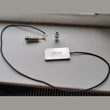 Na fotografii je stříbrný obdélníkový snímač s nápisem KRIVO PICK-UPS. K němu je připojen černý kabel, který končí kovovým jack konektorem. Vedle sní…