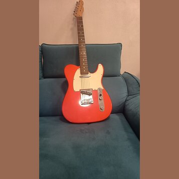 Elektrická kytara s červeným trupem, bílým pickguardem a hnedým hmatníkem. Na hlavě nástroje je viditelné logo Fender. Povrch instrumentu vypadá čist…