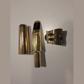 Fotografie zobrazuje zlatý mouton s ligaturem a krytkou. Na moutonu je svisle vyražen nápis "SELMER" a v dolní části nápis "PARIS MADE". Povrch je le…