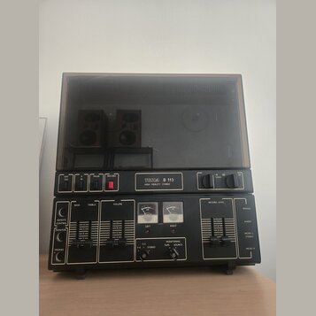 Černý cívkový magnetofon s průhledným plastovým krytem. Na předním panelu je nápis TESLA B 113 a pod ním HIGH FIDELITY STEREO. Zařízení obsahuje něko…