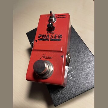 Červený efekt pedál s nápisem PHASER a značkou Rowin. Na horní straně se nachází černý ovládací knoflík s popiskem SPEED a přepínač s nápisy VINTAGE…