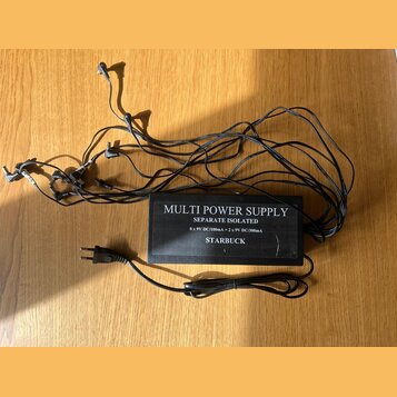 Černý napájecí adaptér s nápisem MULTI POWER SUPPLY, SEPARATE ISOLATED, 8 x 9V DC/100mA + 2 x 9V DC/300mA a STARBUCK. Z jednotky vychází deset černýc…