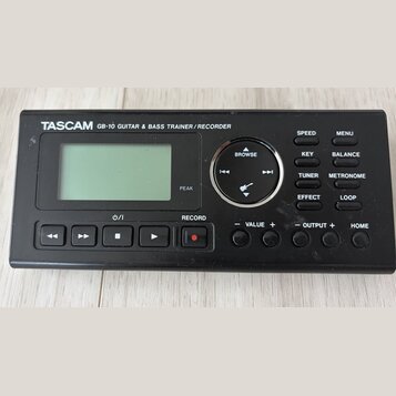 Černý digitální přístroj s nápisem TASCAM GB-10 GUITAR & BASS TRAINER / RECORDER. Zařízení má LCD displej, ovládací kolečko s nápisem BROWSE a řadu t…