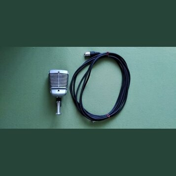 Stříbrný mikrofon v retro stylu s žebrovaným povrchem a kovovým stojankem. K mikrofonu patří černý kabel s konektorem. Povrch mikrofonu vykazuje drob…