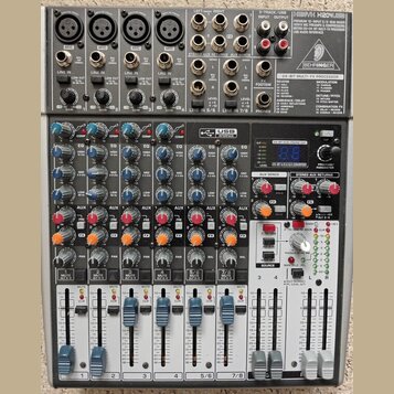 Šedý mixážní pult značky BEHRINGER s modelem XENYX X2022USB. Zařízení obsahuje několik vstupních kanálů s regulačními knoflíky, posuvníky hlasitosti…