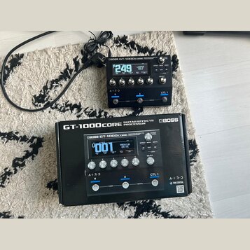 Černý efektový procesor Boss GT-1000CORE s digitálním displejem a několika ovládacími knoflíky. Na zařízení je vidět nápis BOSS GT-1000CORE GUITAR EF…