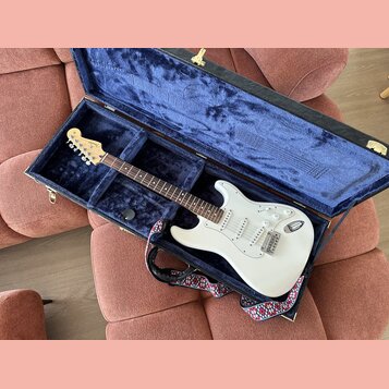 Bílá elektrická kytara typu Stratocaster v otevřeném tvrdém kufru s modrým polstrováním. Na hlavě hmatníku je viditelné logo Fender. Kytara má tři sn…