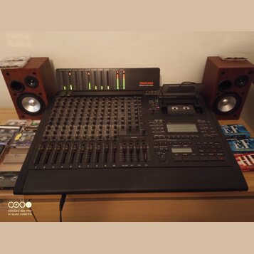 Černý digitální mixážní pult s řadou posuvníků a ovládacích knoflíků. V horní části je umístěn modul s LED indikátory a nápisem TASCAU DIGITAL STUDIO…