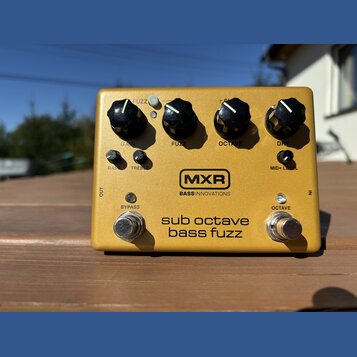 Zlatý efekt pedál s nápisy MXR a BASS INNOVATIONS. Na horní straně jsou čtyři černé oválné knoflíky s popisky GAIN, FUZZ, OCTAVE a DRY. Ve spodní čás…
