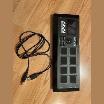 Černý sampler s osmi gumovými pady a displejem. Na přístroji jsou nápisy AKAI professional, MPX8 MOBILE SAMPLE PLAYER a označení 3D. K zařízení je př…