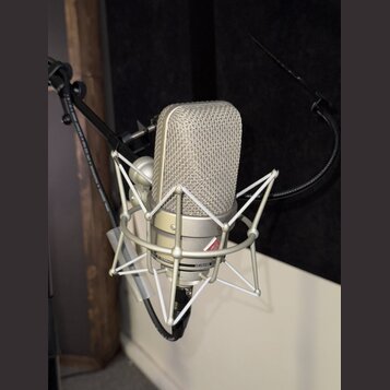 Stříbrný kondenzátorový mikrofon v šokabsorbujícím držáku. Na těle mikrofonu je viditelný nápis NEUMANN a text MADE IN GERMANY. Povrch mikrofonu i dr…