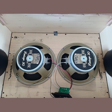 Dva reproduktory značky CELESTION s nápisy LOUDSPEAKERS, G12 a Vintage 30. Reproduktory mají béžové rámy a jsou umístěny v dřevěné bedně. Mezi nimi j…