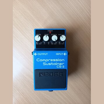 Modrý efekt pedál s nápisy Compression Sustainer a CS-3. Na horní části jsou čtyři bílé ovládací knoflíky s popisky LEVEL, TONE, ATTACK a SUSTAIN, če…