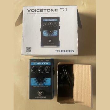 Fotografie zobrazuje efekt pedál VoiceTone C1 značky TC HELICON v původním kartonu s přibaleným napájecím adaptérem. Na zařízení je vidět nápis TC HE…
