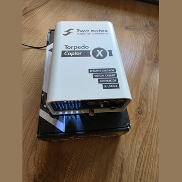 Bíle provedené zařízení s kovovým šasi a černými nápisy. Na horní části je logo a text Two notes AUDIO ENGINEERING, dále označení Torpedo Captor X a…
