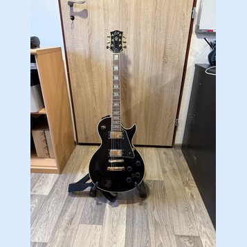 Černá elektrická kytara s gold hardwarem umístěná na stojanu. Na hlavě hmatníku je viditelné logo Epiphone. Kytara má dva humbuckery, čtyři ovládací…