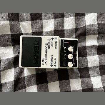 Bílý efekt pedál s černým stupňovitým šlapadlem. Na těle jsou nápisy BOSS, Noise Suppressor NS-2 a označení konektorů OUTPUT, SEND, RETURN, INPUT. Ov…