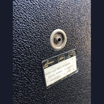 Detail černého povrchu basového komba s texturou. Na povrchu je kovový konektor a štítek s nápisy: „ELECTRONICS CORP“, logo „Peavey“ a adresa „711 A…