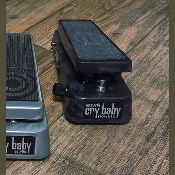 Dvě wah pedály umístěné na dřevěném povrchu. Pravý pedál má černý maskáčový vzor a nápis DIME cry baby FROM HELL. Levý pedál je stříbrný s nápisem cr…
