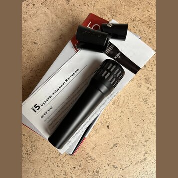 Černý dynamický mikrofon s mřížkou, položený na bílé krabici. Na krabici je nápis i5 dynamic Instrument Microphone a sekce OVERVIEW s popisem produkt…