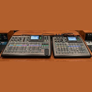Dvě digitální mixovací konzole černé barvy umístěné vedle sebe na dřevěném povrchu. Na levé konzoli je v horní části viditelné logo a nápis X32. Obě…
