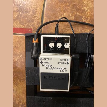Bílý efekt pedál značky BOSS s nápisem Noise Suppressor NS-2. Zařízení má tři ovládací knoflíky s popisky REDUCTION, THRESHOLD a DECAY, přepínač MODE…
