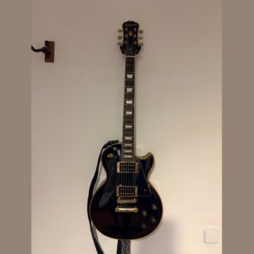 Černá elektrická kytara typu Les Paul se zlatým hardwarem a bílými blokovými vložkami na hmatníku. Na hlavě je viditelné logo Epiphone. Kytara je zav…