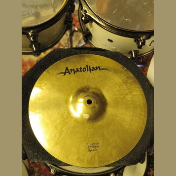 Zlatý cymbál typu splash položený na černém texturovaném podkladu. V horní části je černé logo „Anatolian“. Dole je nápis „Ultimate 12"/30cm Splash“.…